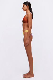 Woman wearing rust-colored racerback sports bra with yellow and purple colorblock bikini bottoms, shown in profile view.
