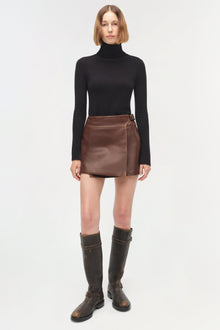 Brown leather mini skirt with wrap-front design and buckle closure, paired with black turtleneck and boots.