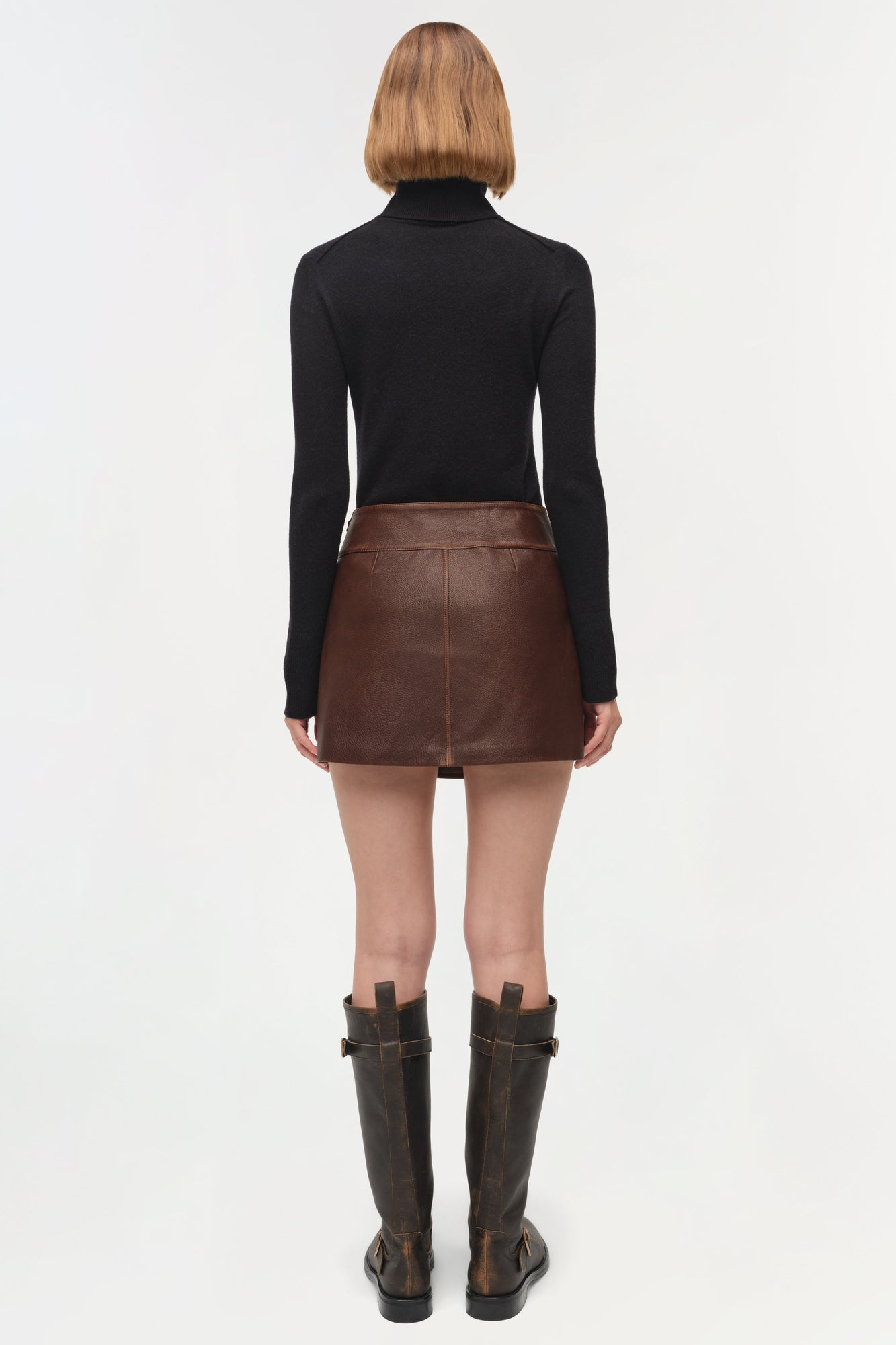 Brown leather mini skirt with front seam detail, worn with black turtleneck and dark boots, back view.