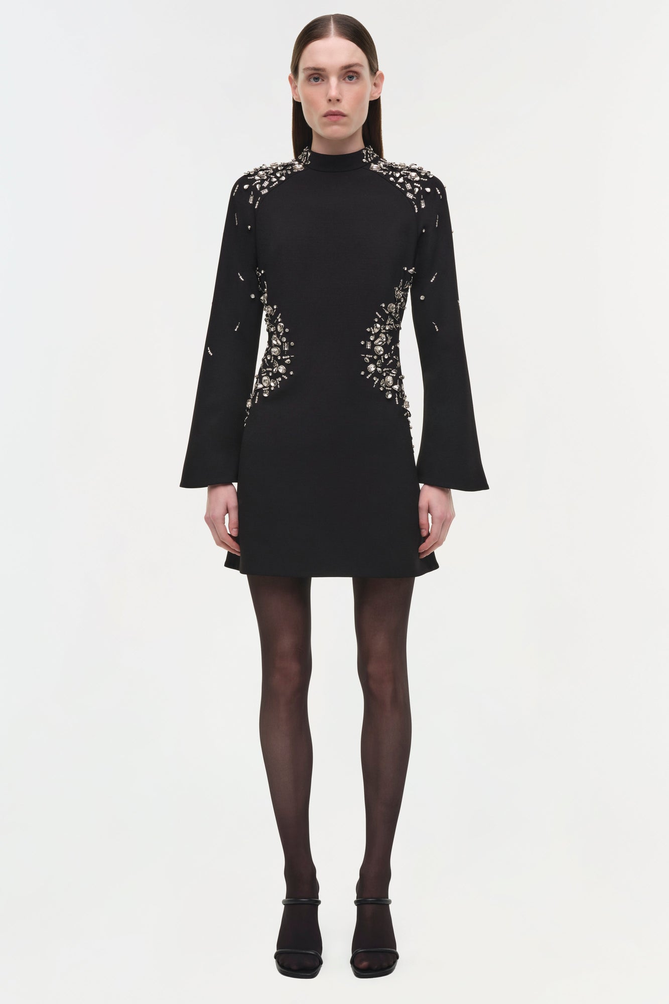 Black mini dress with silver crystal embellishments on shoulders, sides, and sleeves; long flared sleeves and high neckline.
