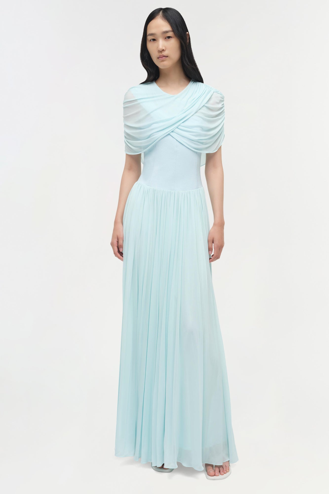 Light blue draped jersey maxi dress with pleated skirt and draped cape overlay across shoulders.
