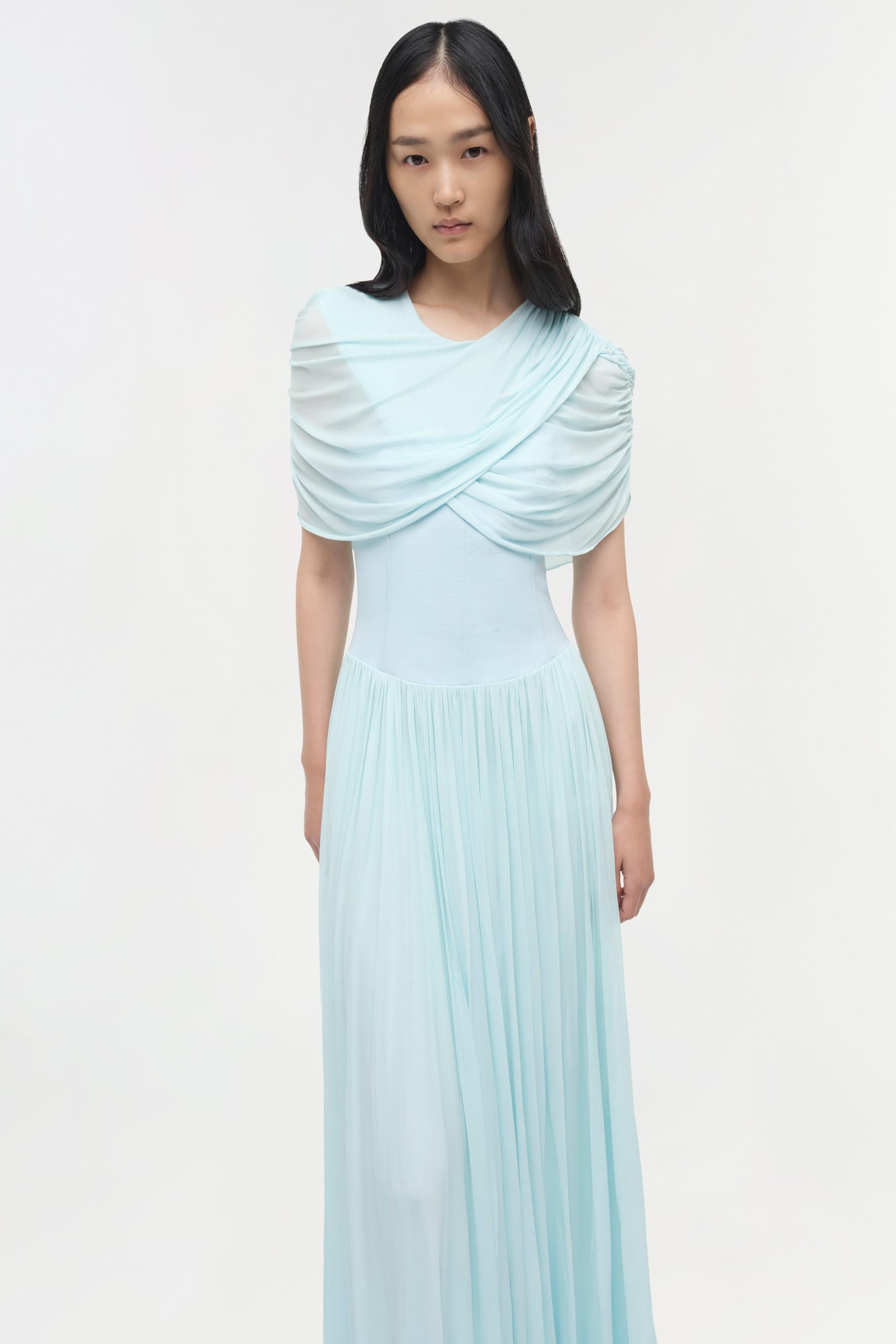 Light blue jersey maxi dress with draped cape overlay and pleated skirt.