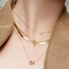 Lifestyle Model Close-up of a woman wearing a camel-colored top and four layered gold necklaces: a delicate single diamond pendant, a flat herringbone chain, a paperclip chain with a diamond 'S' pendant, and a longer chain with interlocking diamond rings