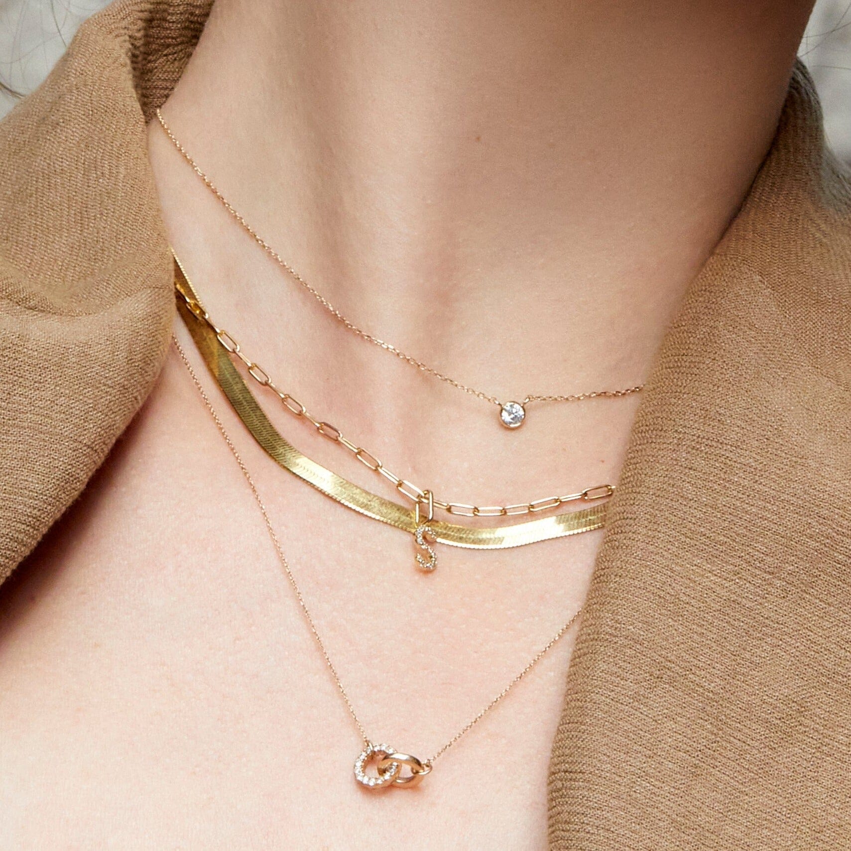 Lifestyle Model Close-up of a woman wearing a camel-colored top and four layered gold necklaces: a delicate single diamond pendant, a flat herringbone chain, a paperclip chain with a diamond 'S' pendant, and a longer chain with interlocking diamond rings