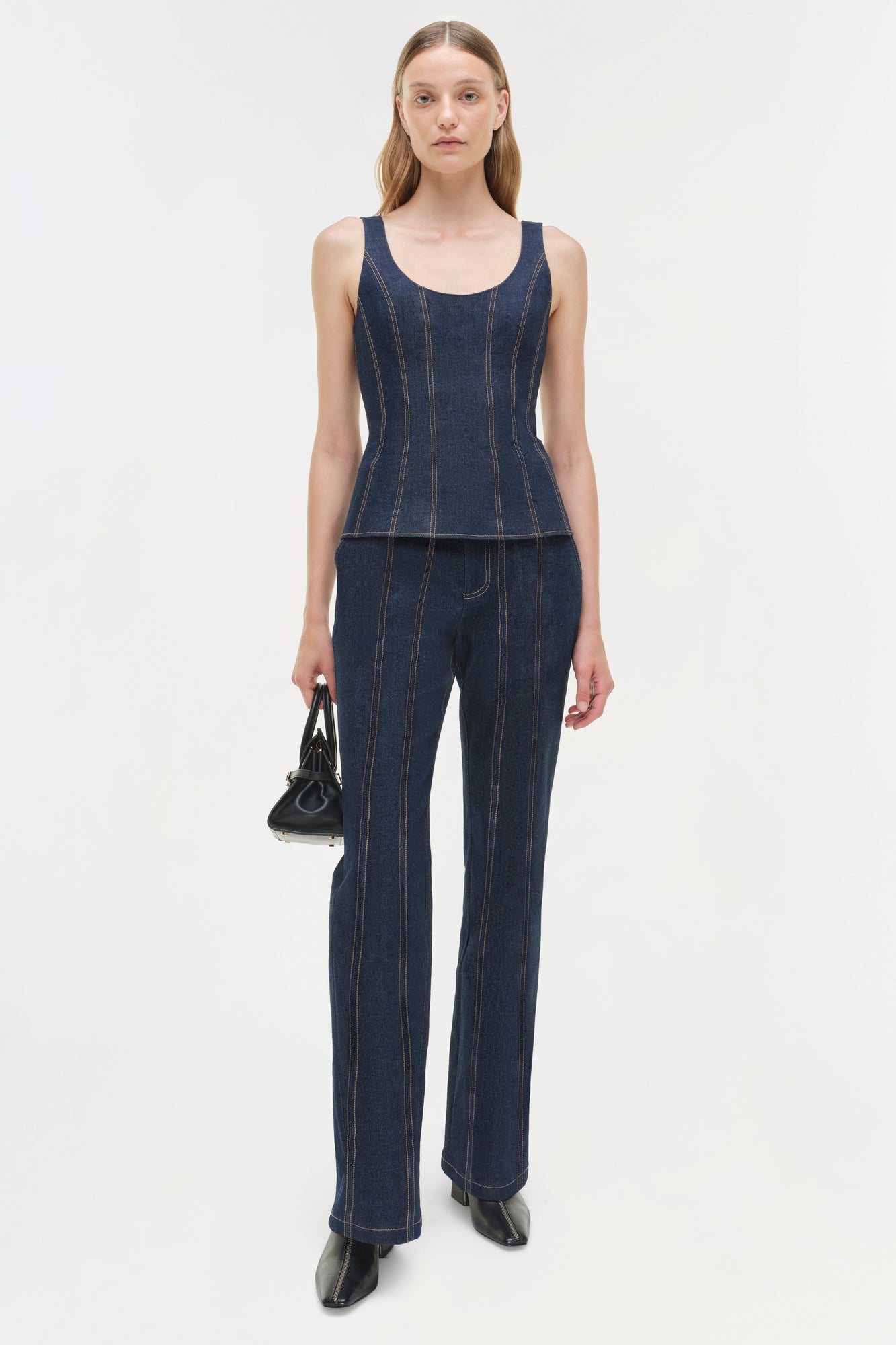 Woman wearing dark blue denim trousers with vertical stitching and matching sleeveless top, holding black handbag.
