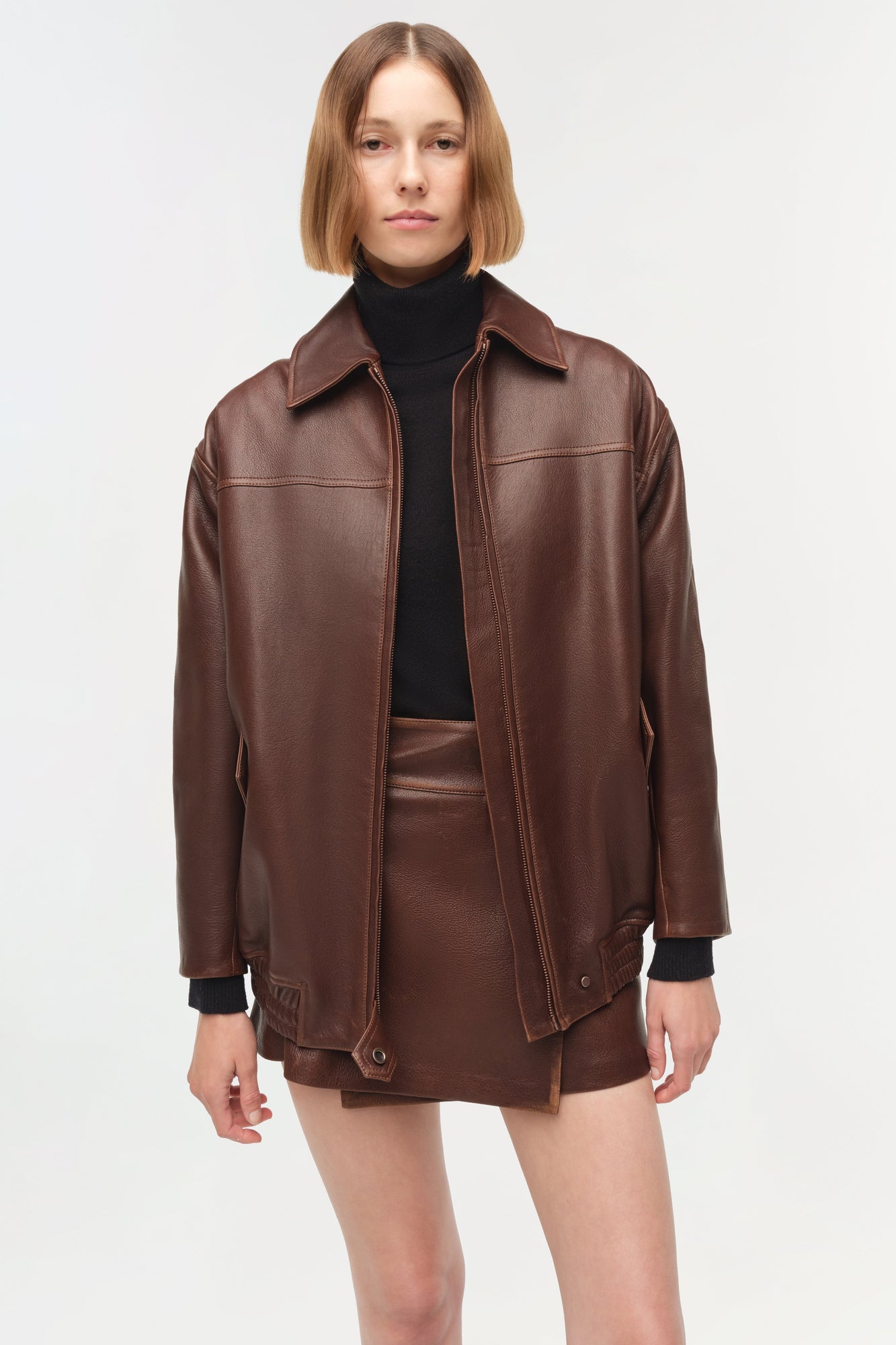 Brown leather oversized jacket with wide collar, full-length sleeves, and front zip closure worn over black turtleneck.