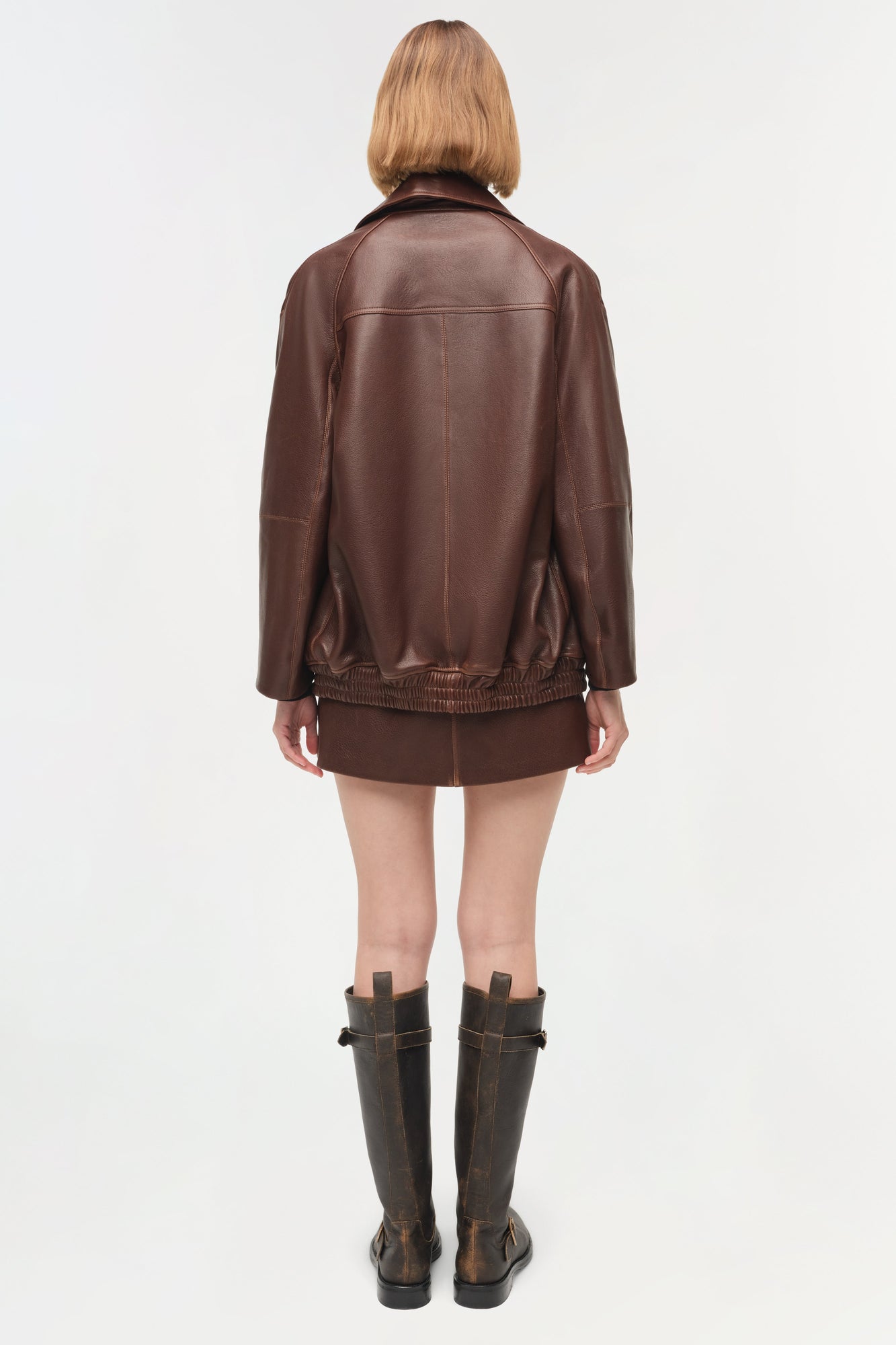 Brown leather jacket with ribbed hem and collar, shown from back on model wearing matching shorts and dark boots.