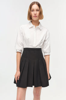 Woman wearing white button-up shirt with three-quarter sleeves over black pleated mini skirt.