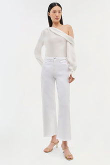 White off-shoulder long-sleeve top with draped neckline on model wearing white cropped jeans.