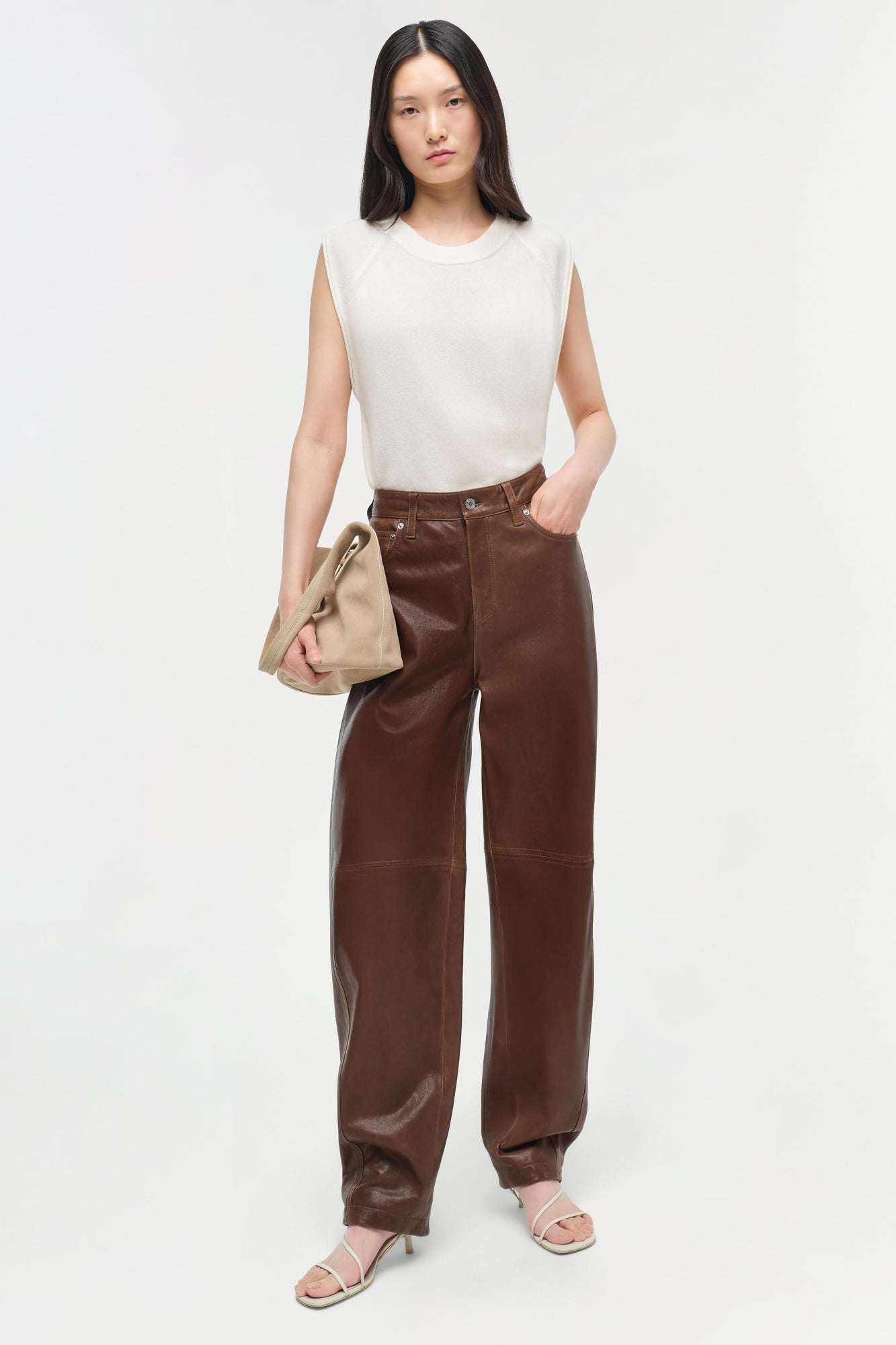 Woman wearing a cream sleeveless cashmere vest with brown leather pants and holding a beige bag.