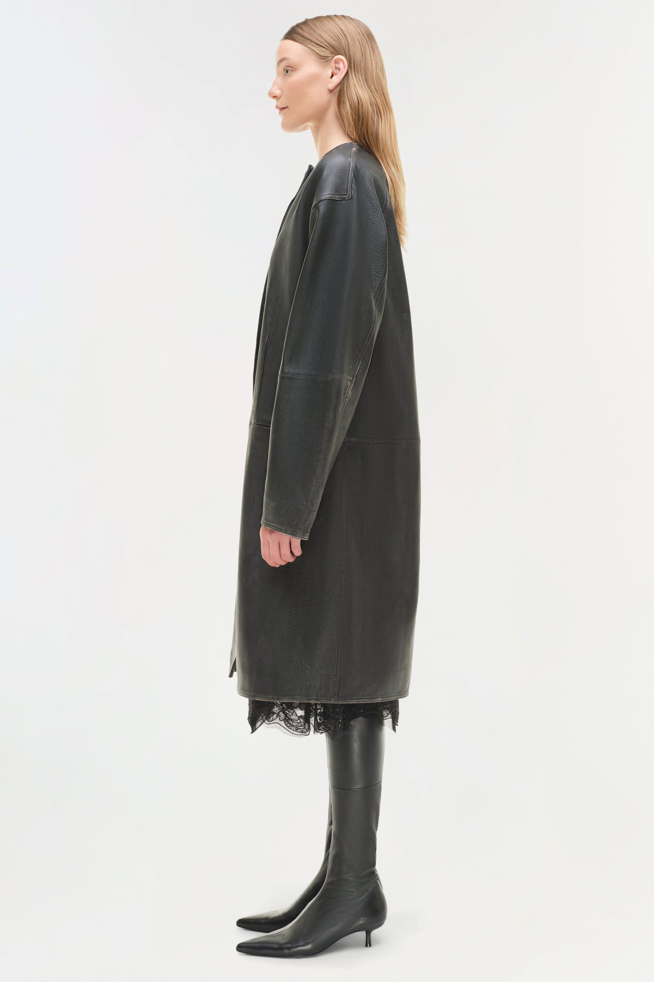 Woman wearing a charcoal leather coat with long sleeves, lace hem detail, and black leather knee-high boots, shown in profile