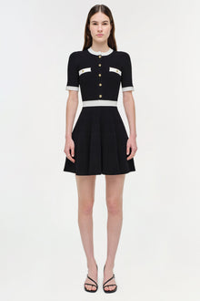 Black ribbed knit mini dress with white trim, gold button front closure, and pleated skirt.