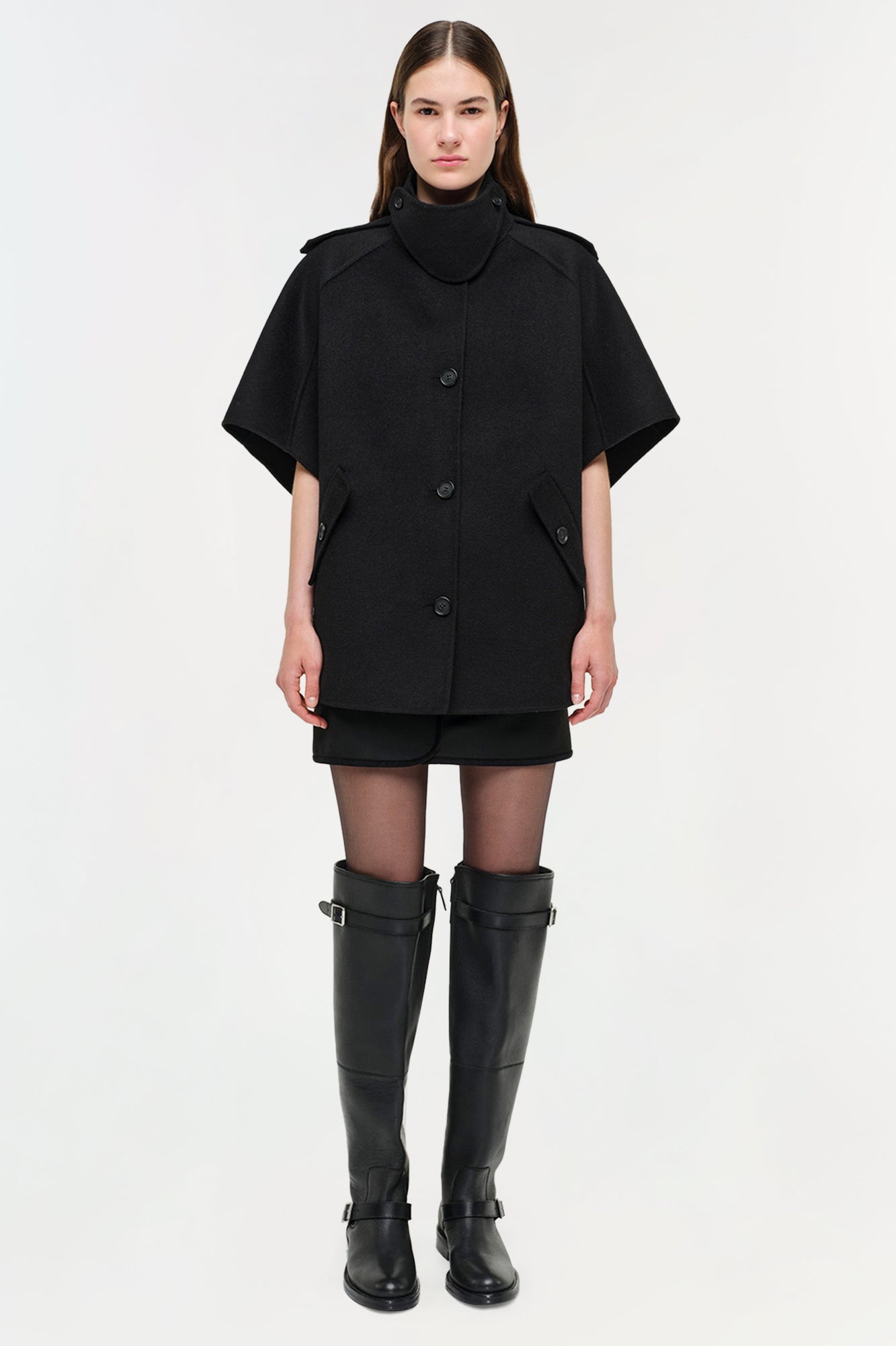 Black wool cape jacket with button front, high neckline, and short sleeves worn with black boots.