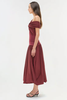 Woman wearing burgundy off-shoulder top with draped neckline and full midi skirt, side view.