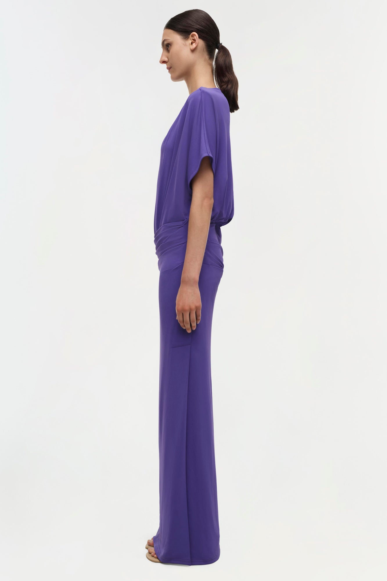 Purple floor-length gown with draped short sleeves and fitted waist, shown from side profile on model.