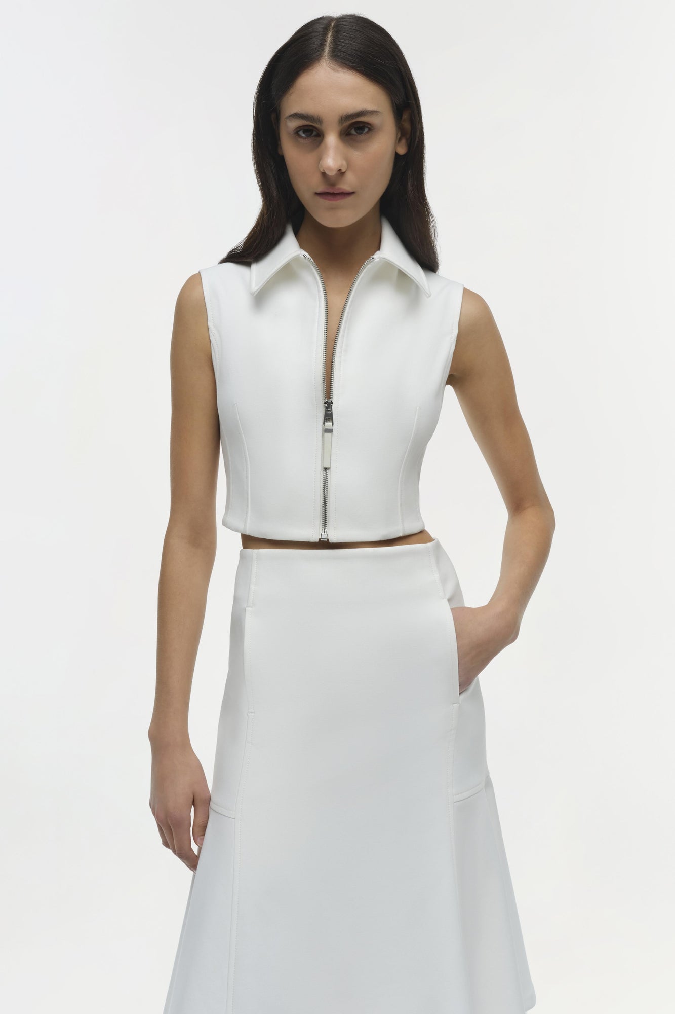 White sleeveless zip-up vest with collared neckline, worn over matching white skirt with side pockets.