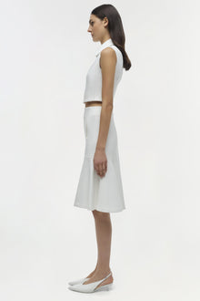 Woman wearing white sleeveless collared crop top with matching knee-length skirt and white kitten heels, shown in profile vie