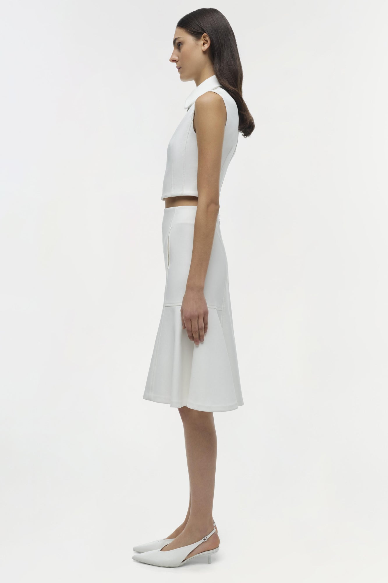 Woman wearing white sleeveless collared crop top with matching knee-length skirt and white kitten heels, shown in profile vie