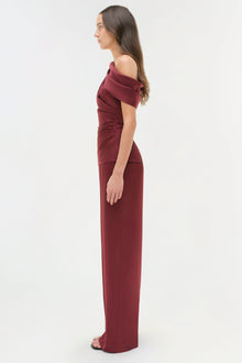 Woman wearing a burgundy draped satin gown with off-shoulder neckline and floor-length skirt.