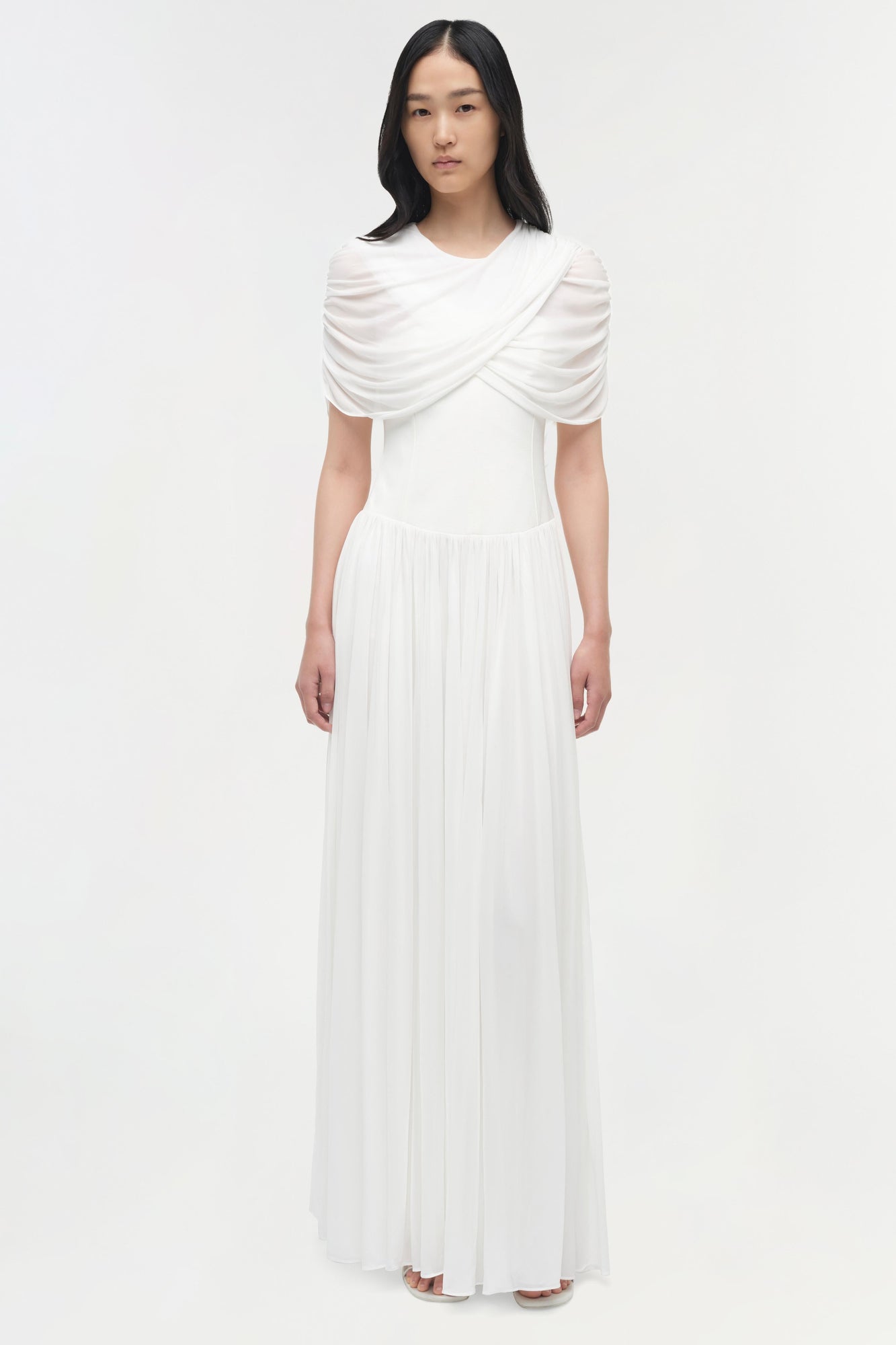 White jersey maxi dress with draped cape overlay and pleated skirt.