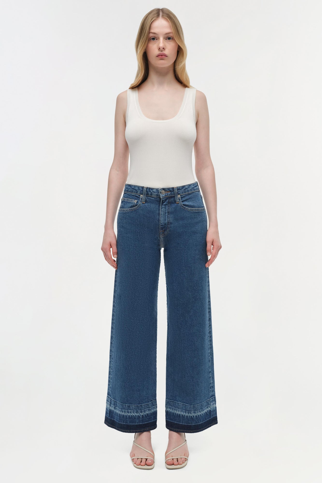 Woman wearing white sleeveless tank top and blue wide-leg cropped jeans with darker hem.