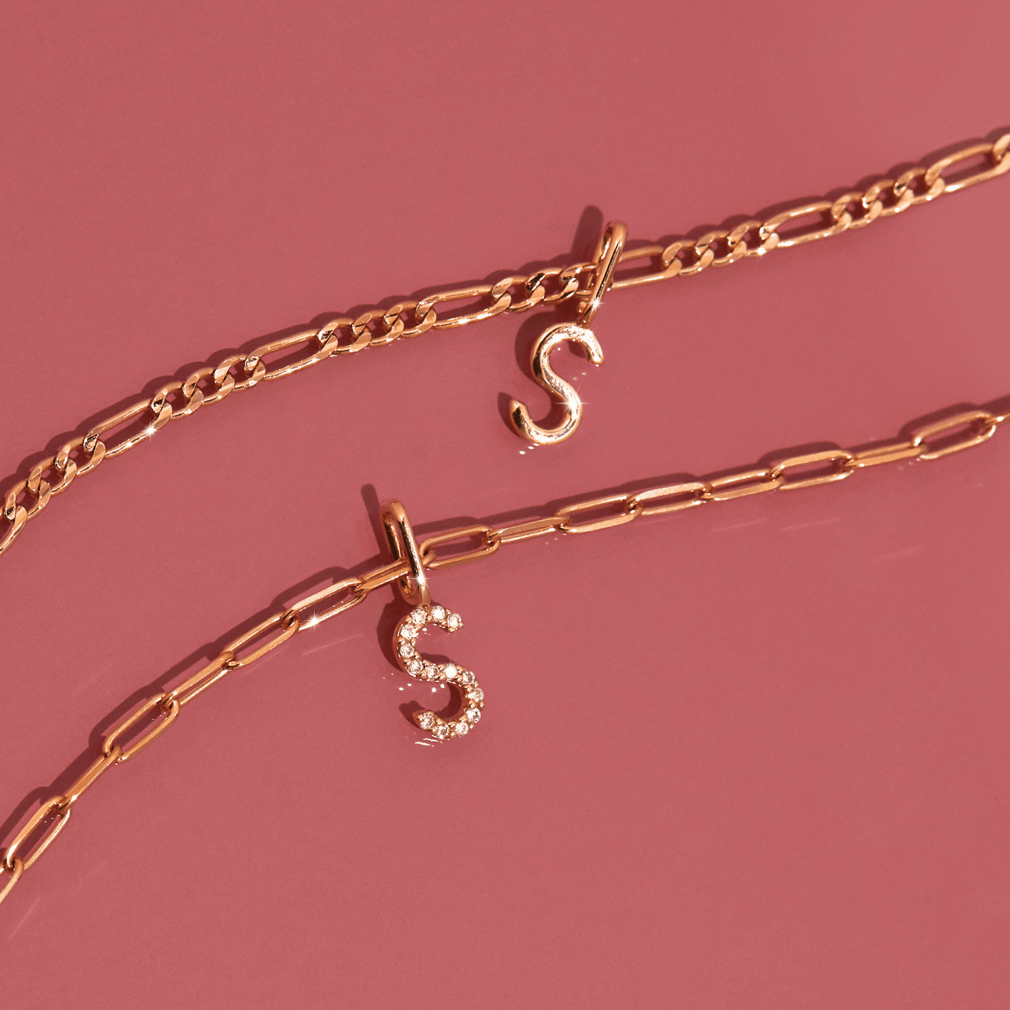 Flatlay Model Close-up of two gold chain necklaces with 'S' pendantsâ€”one solid gold and one diamond-encrustedâ€”resting against a pink background