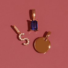 Flatlay Model Close-up of three gold pendantsâ€”a rectangular blue gemstone, a diamond-encrusted 'S' initial, and a solid gold round discâ€”arranged on a pink background