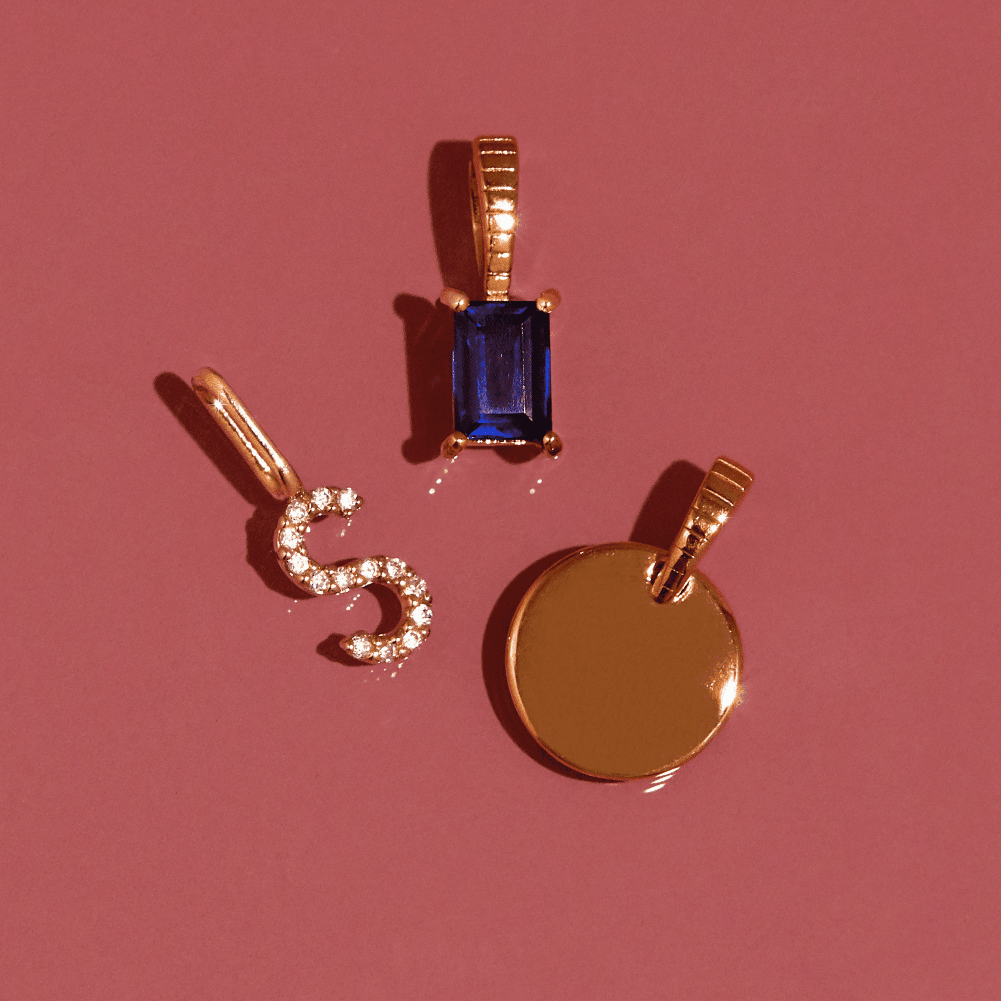 Flatlay Model Close-up of three gold pendantsâ€”a rectangular blue gemstone, a diamond-encrusted 'S' initial, and a solid gold round discâ€”arranged on a pink background