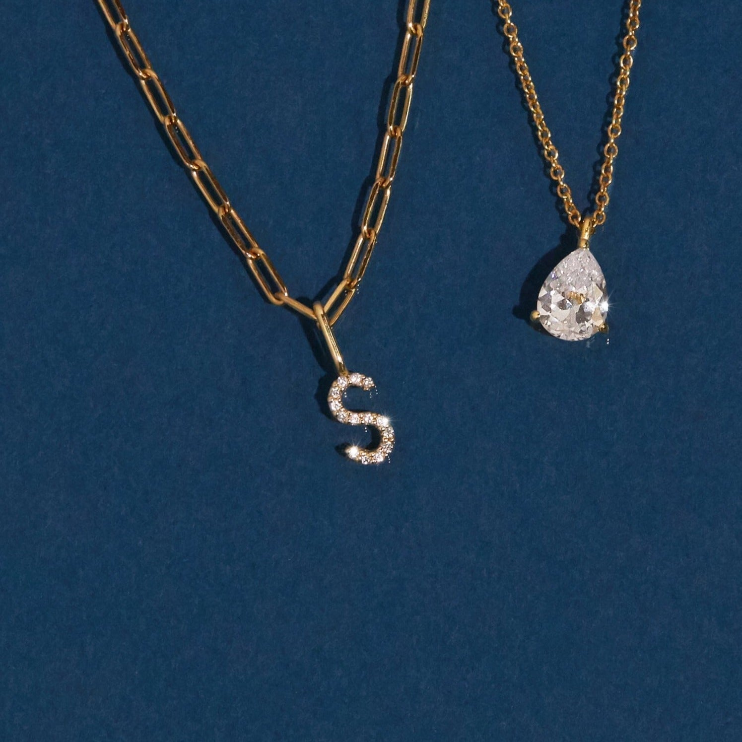 Flatlay Model Close-up of two gold necklaces resting against a dark blue backgroundâ€”one featuring a diamond-encrusted 'S' pendant and the other a pear-shaped clear gemstone pendant