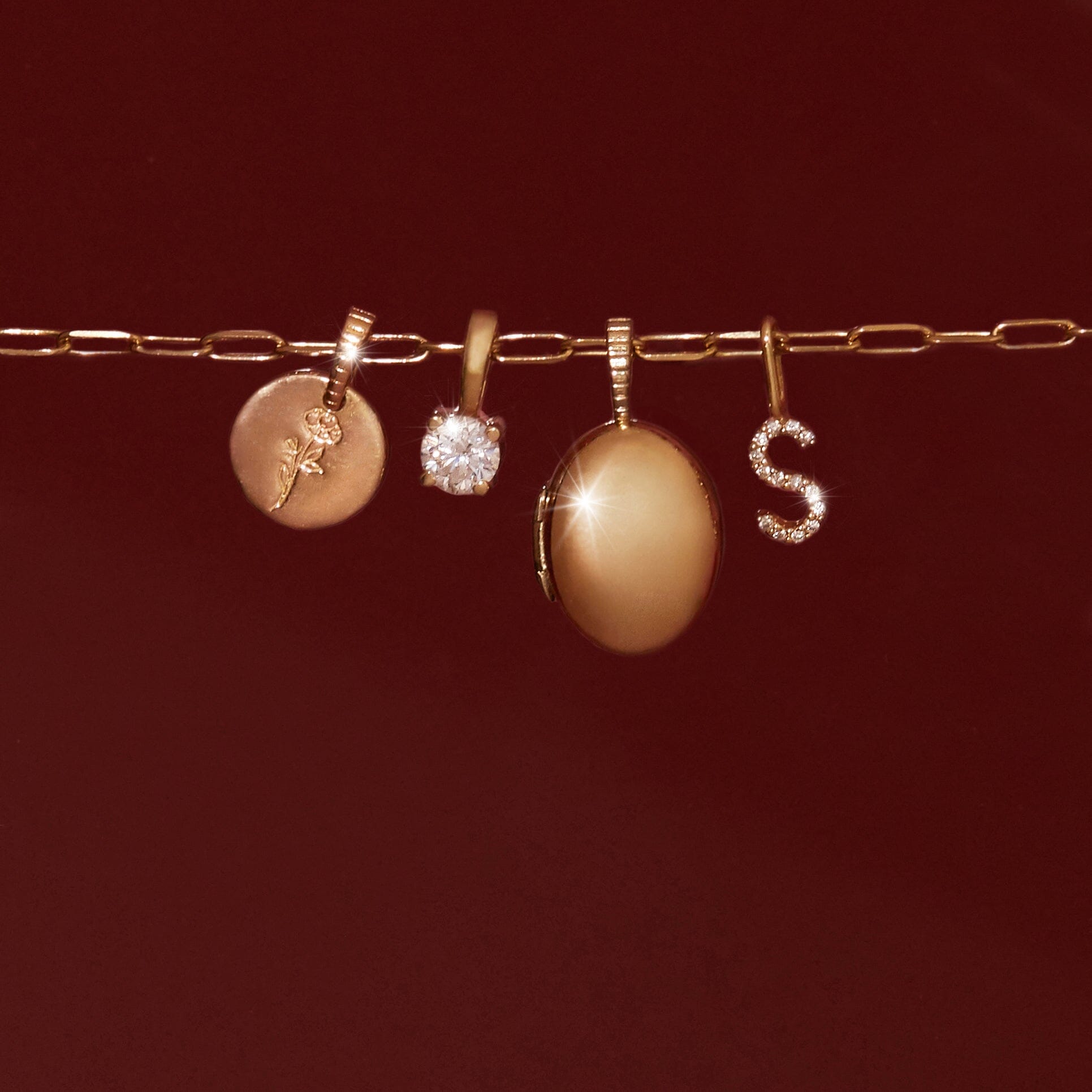 Flatlay Model Close-up of a gold paperclip chain displaying four pendantsâ€”a gold disc with a rose engraving, a round clear gemstone, a smooth gold oval locket, and a diamond-encrusted 'S' initialâ€”resting against a dark red background