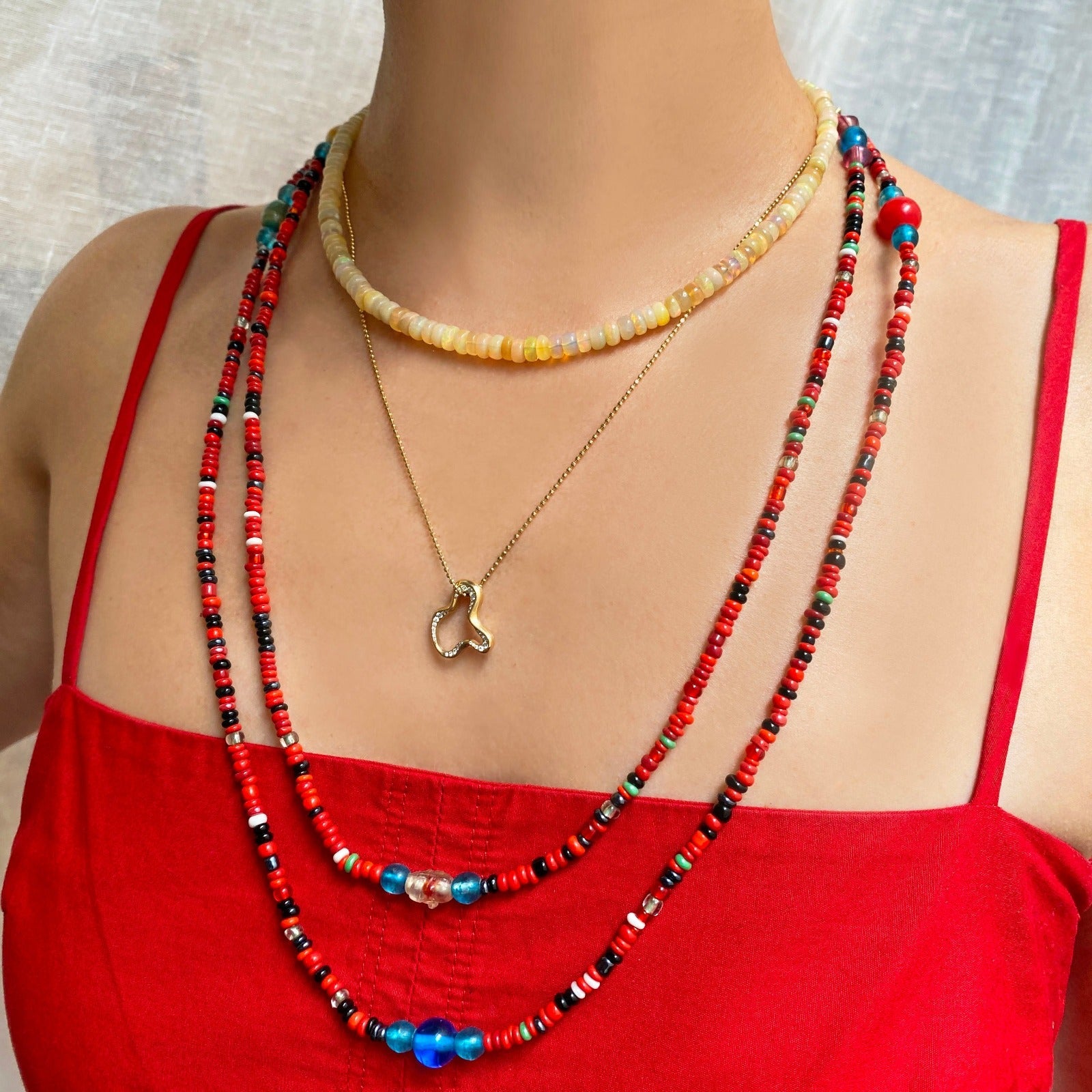 Shimmering beaded necklace made of smooth opals in shades of yellow, tan and white on a gold linking ovals clasp on a neck layered with a chain and charm along with two red beaded neckalces