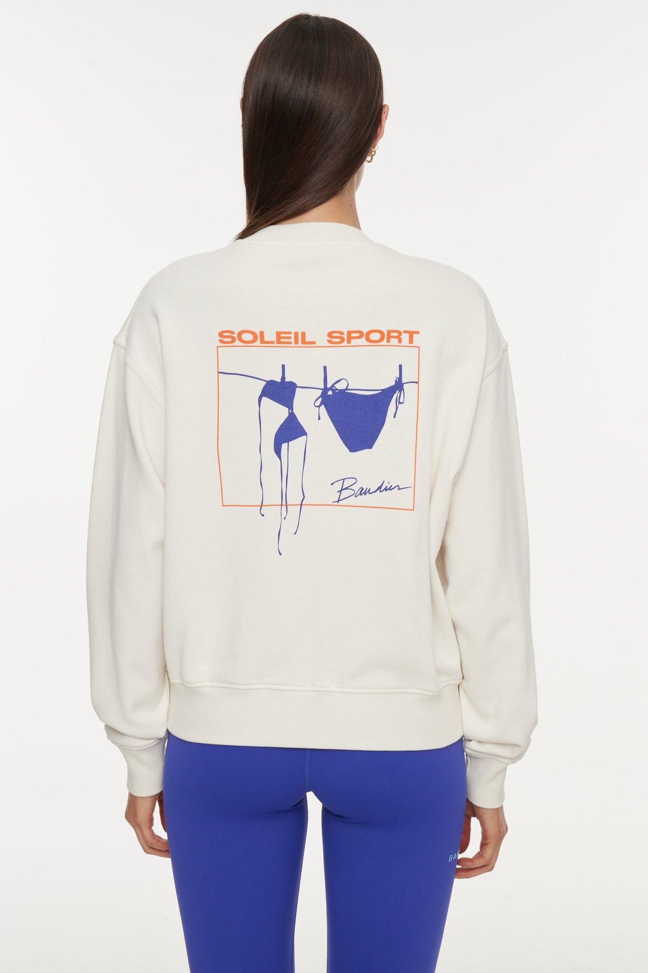 Model wears a white crewneck sweatshirt with logo on back that reads "Soleil Sport" and underneath is a square orange border containing a blue bikini hanging to dry on a clothesline. 