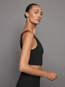 Model wears a black, longline sports bra with a deep V-neck front and an open V-shaped back, made from sleek, smooth fabric.

