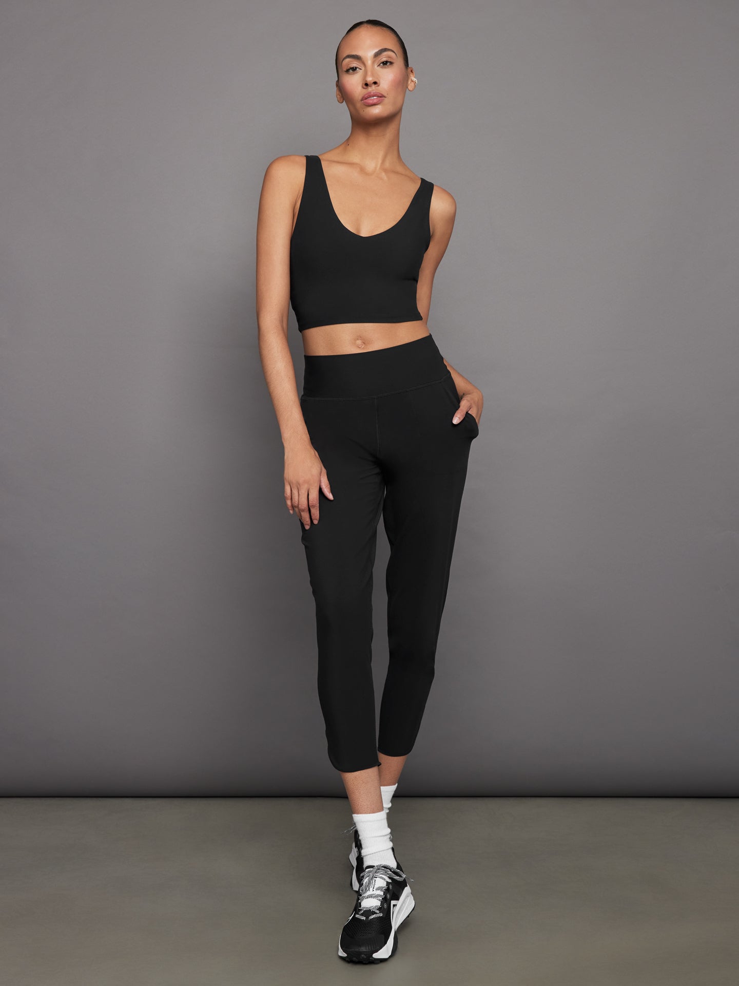 Model wears a black, longline sports bra with a deep V-neck front and an open V-shaped back, made from sleek, smooth fabric.
