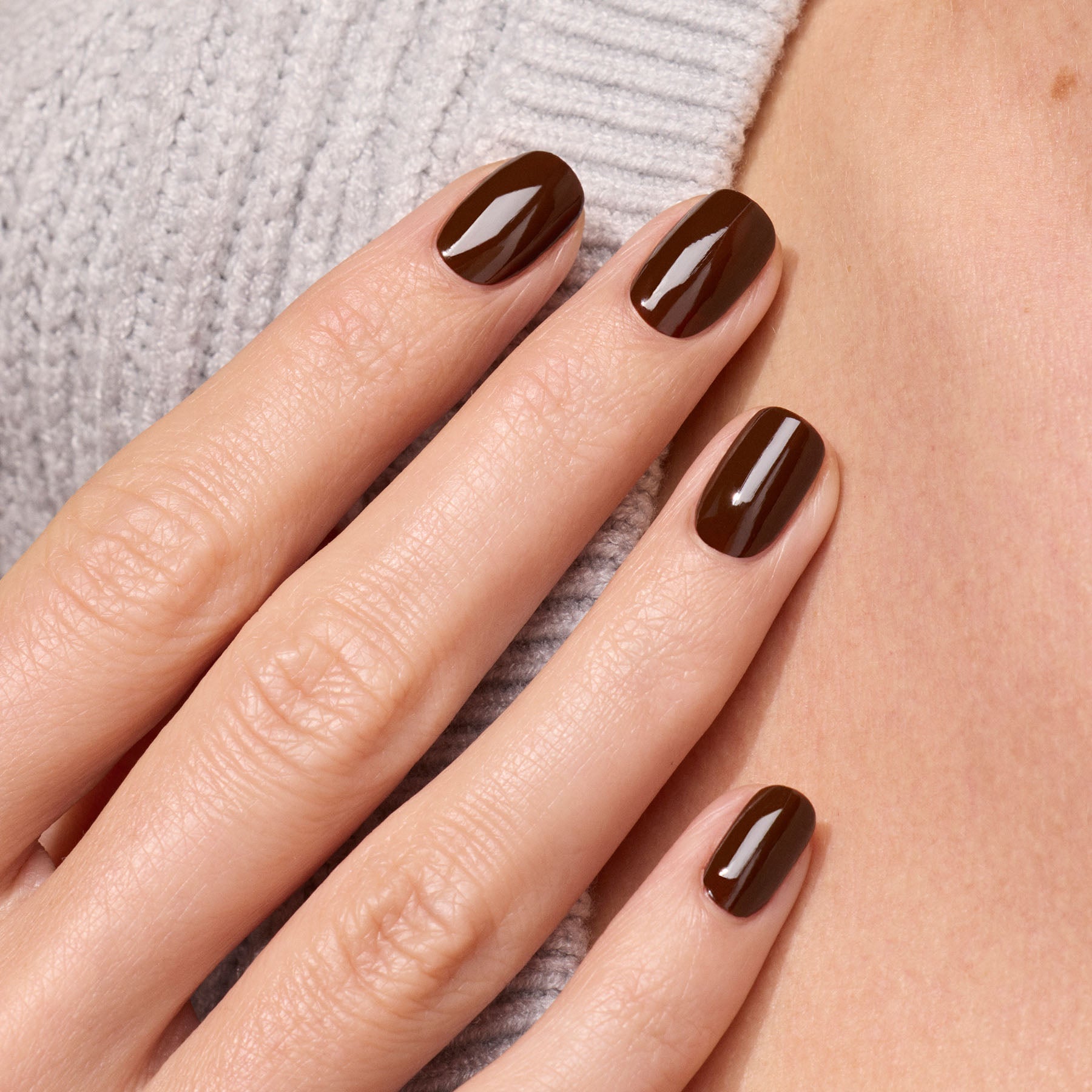 A close-up of a hand with neatly manicured nails painted in Manucurist's Clove, a glossy chocolate brown polish, gently resting on a light gray ribbed sweater.