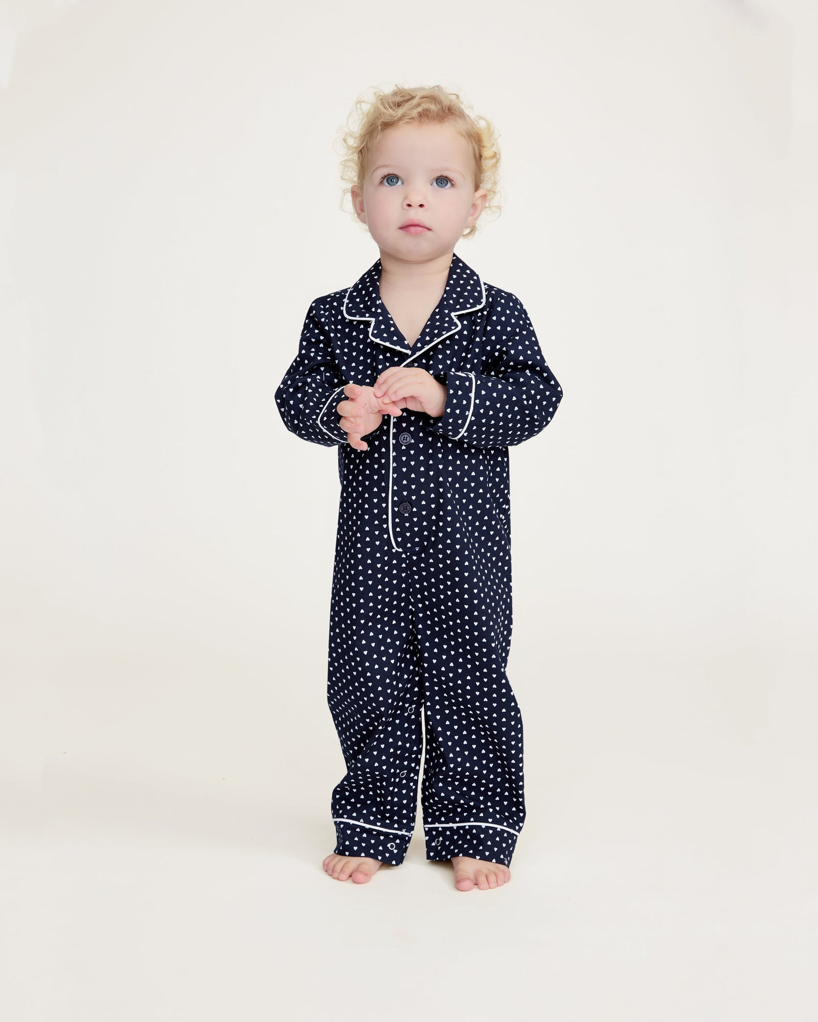 The Baby's Twill Classic Romper in Midnight Hearts is a navy blue onesie with long sleeves and legs, featuring a white heart pattern and white piping on the collar, cuffs, and front.