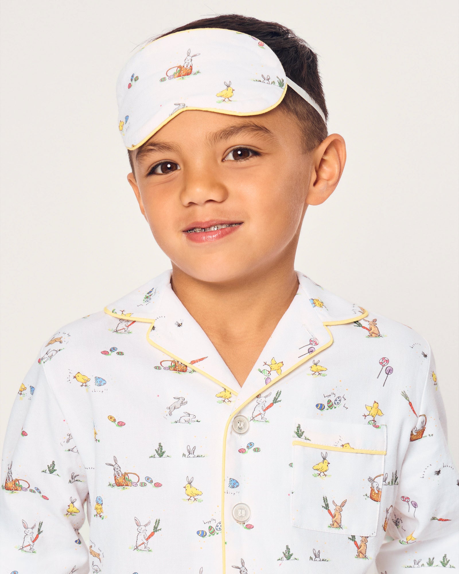 A young boy wears a Kid's Twill Sleep Mask in Easter Frolic, featuring colorful animal and nature prints, along with matching white pajamas. He smiles softly against a plain, light background.