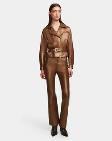 A woman wears the Rudsak ARIELLE - 8124052 Antique Bronze metallic brown relaxed-fit jacket with a belted waist and matching high-waisted leather pants, paired with pointed-toe boots. She has straight hair and a neutral expression against a plain background.