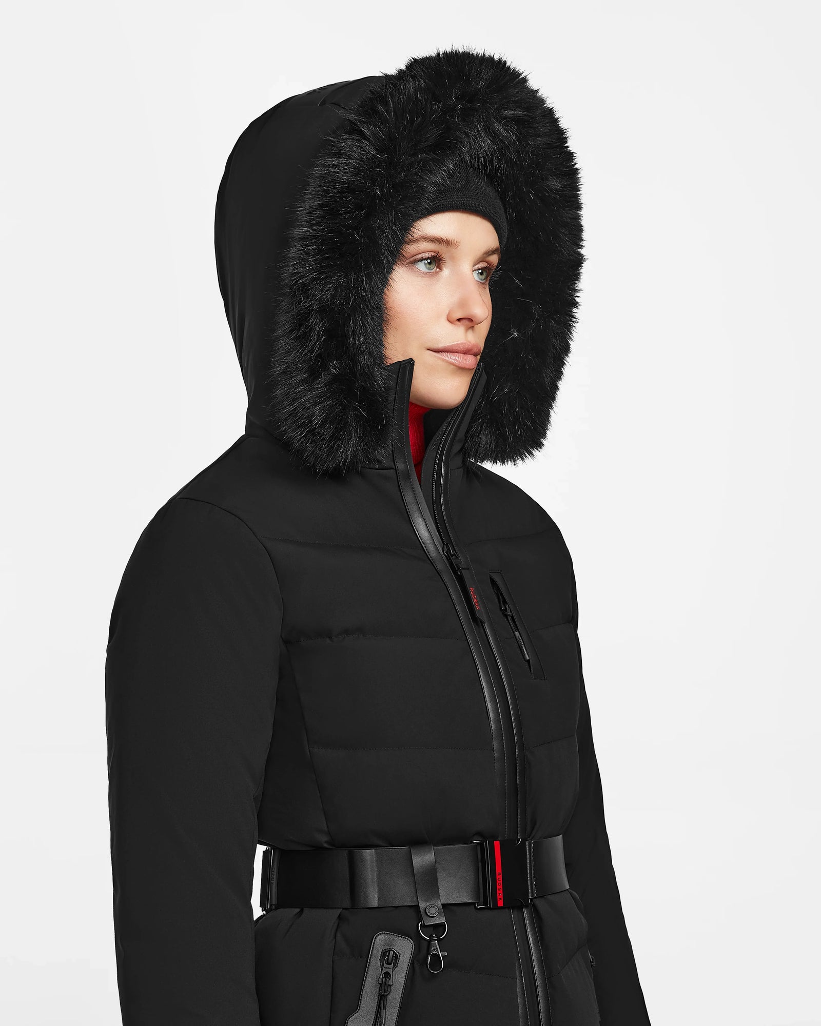 A woman models the Rudsak ALAIA - 8124816 Black, a belted winter coat with a black faux fur-trimmed hood and ski-inspired features, set against a plain white background.