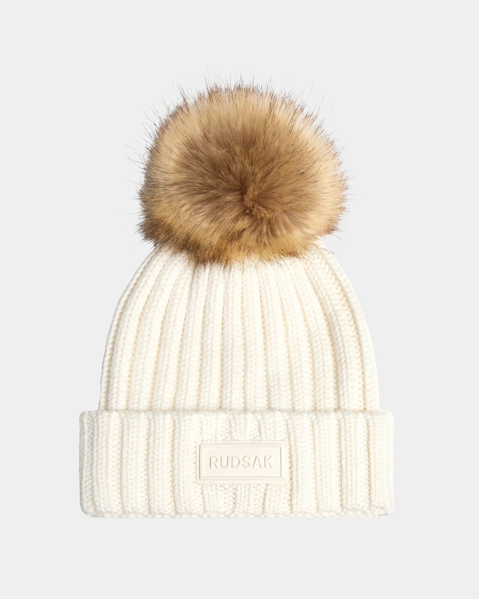 The ANISA - L423500 Optic White by Rudsak is a women's white ribbed knit beanie with a folded brim, brown faux fur pom pom, and a rectangular Rudsak patch on the front. Set against a plain light gray background.