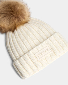 The ANISA - L423500 Optic White by Rudsak is a cream ribbed knit beanie with a folded cuff, large brown faux fur pom pom, and a rectangular patch featuring the Rudsak brand name—perfect for elevating your winter style.
