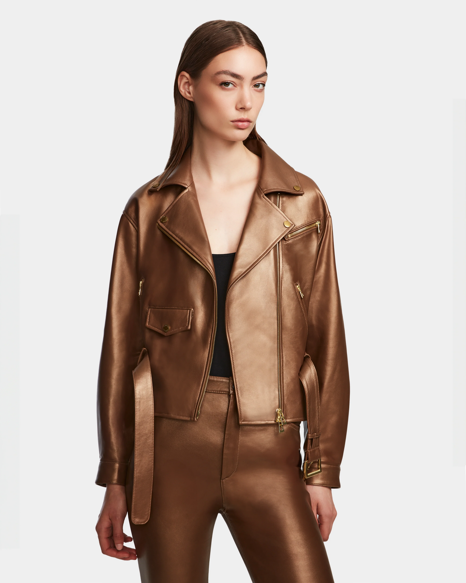 A woman with straight brown hair wears the Rudsak ARIELLE - 8124052 Antique Bronze lamb leather biker jacket and matching pants over a black top, standing against a plain white background.