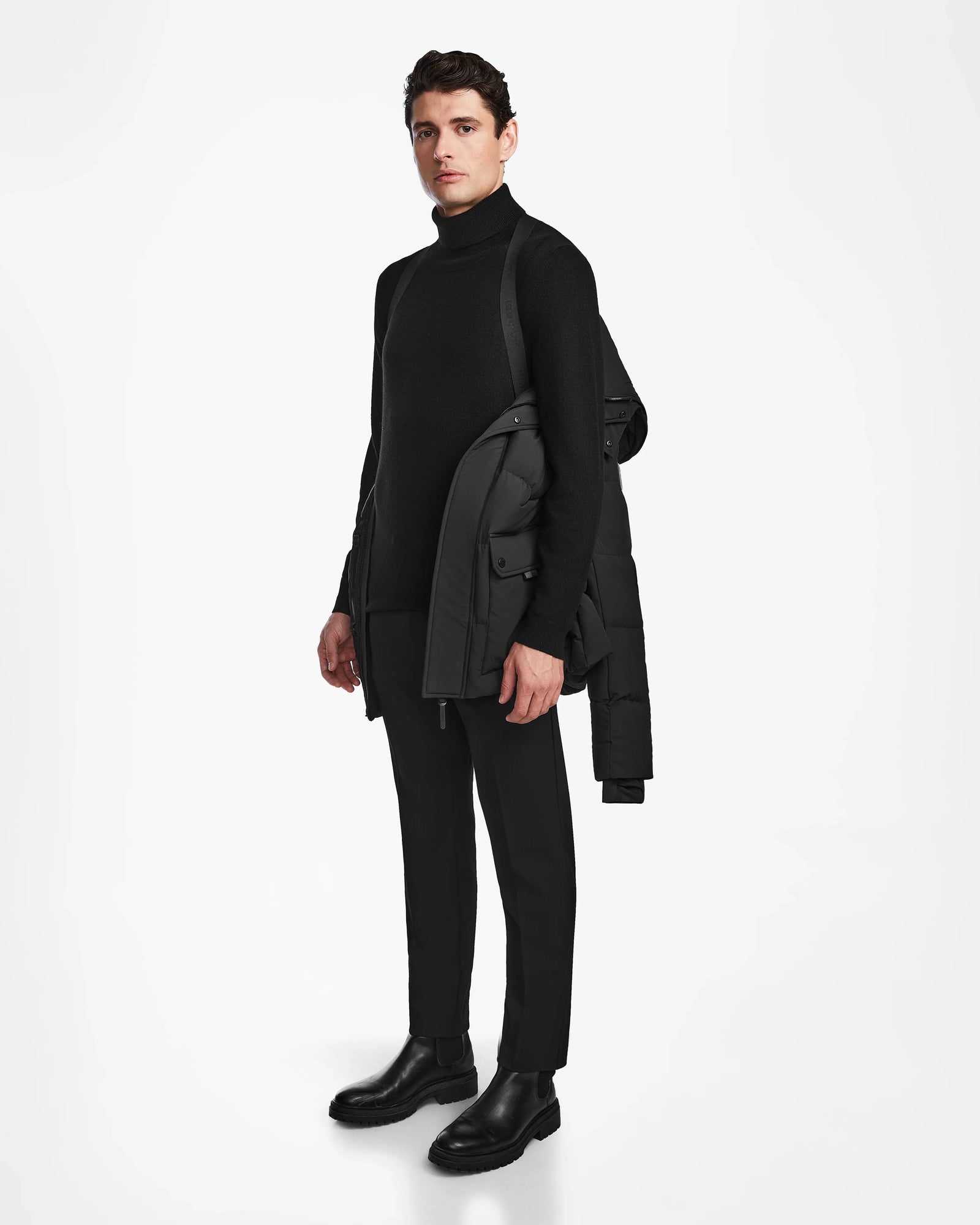 A man wears the Rudsak CARLOS - M124509 Black quilted parka draped over one shoulder, paired with a black turtleneck, trousers, and boots. He stands against a white background, facing slightly left and looking forward.