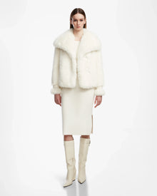 A woman with straight brown hair and a neutral expression stands against a white background, wearing the Rudsak CHLOE - L124564 Iceland faux fur jacket, a cream midi dress with side slit, and knee-high white heeled boots.