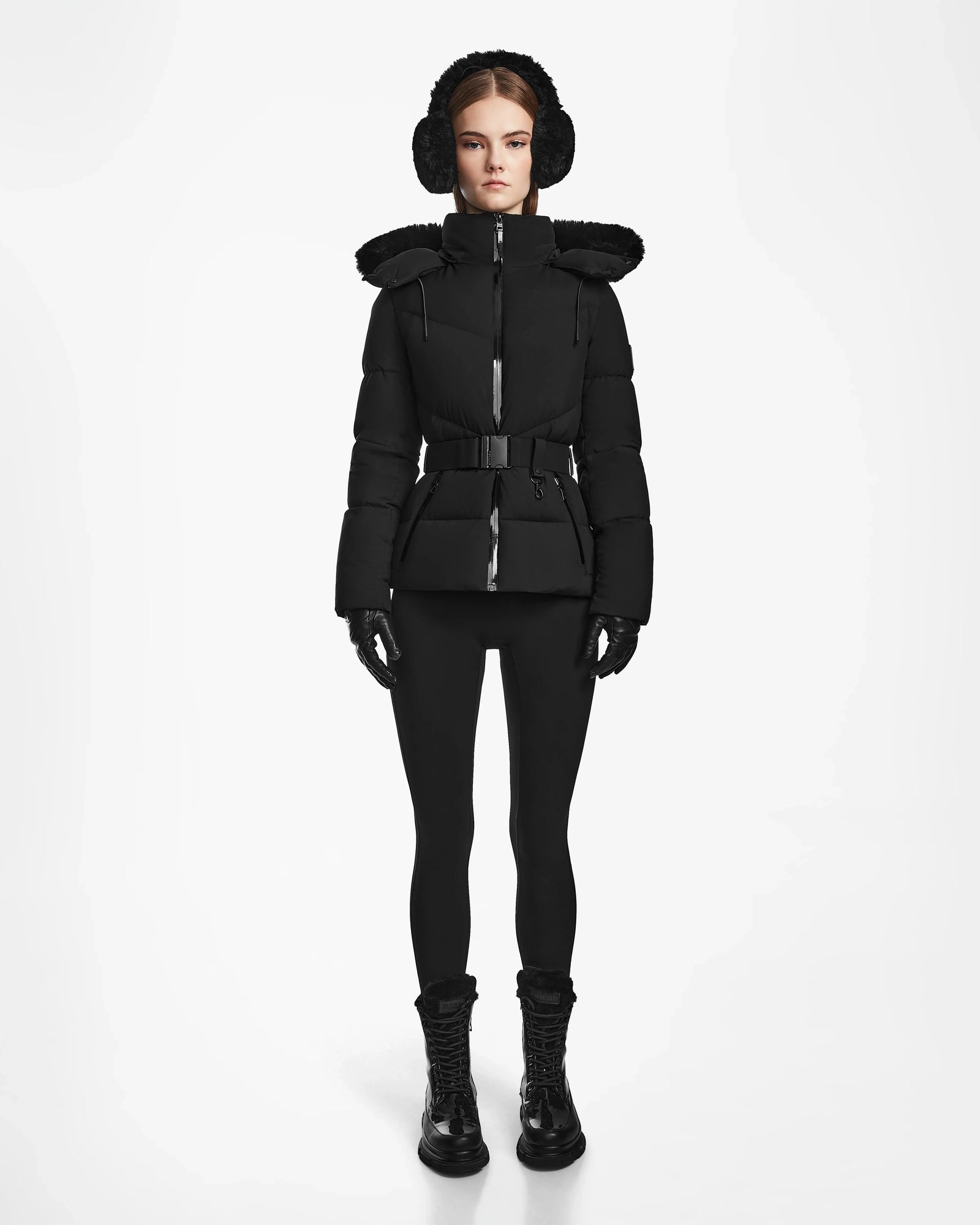 A woman faces forward against a white background, wearing the Rudsak CHRISTELLE M - L123525 Black quilted down puffer with a fur-lined hood, matching earmuffs, gloves, leggings, and lace-up boots.