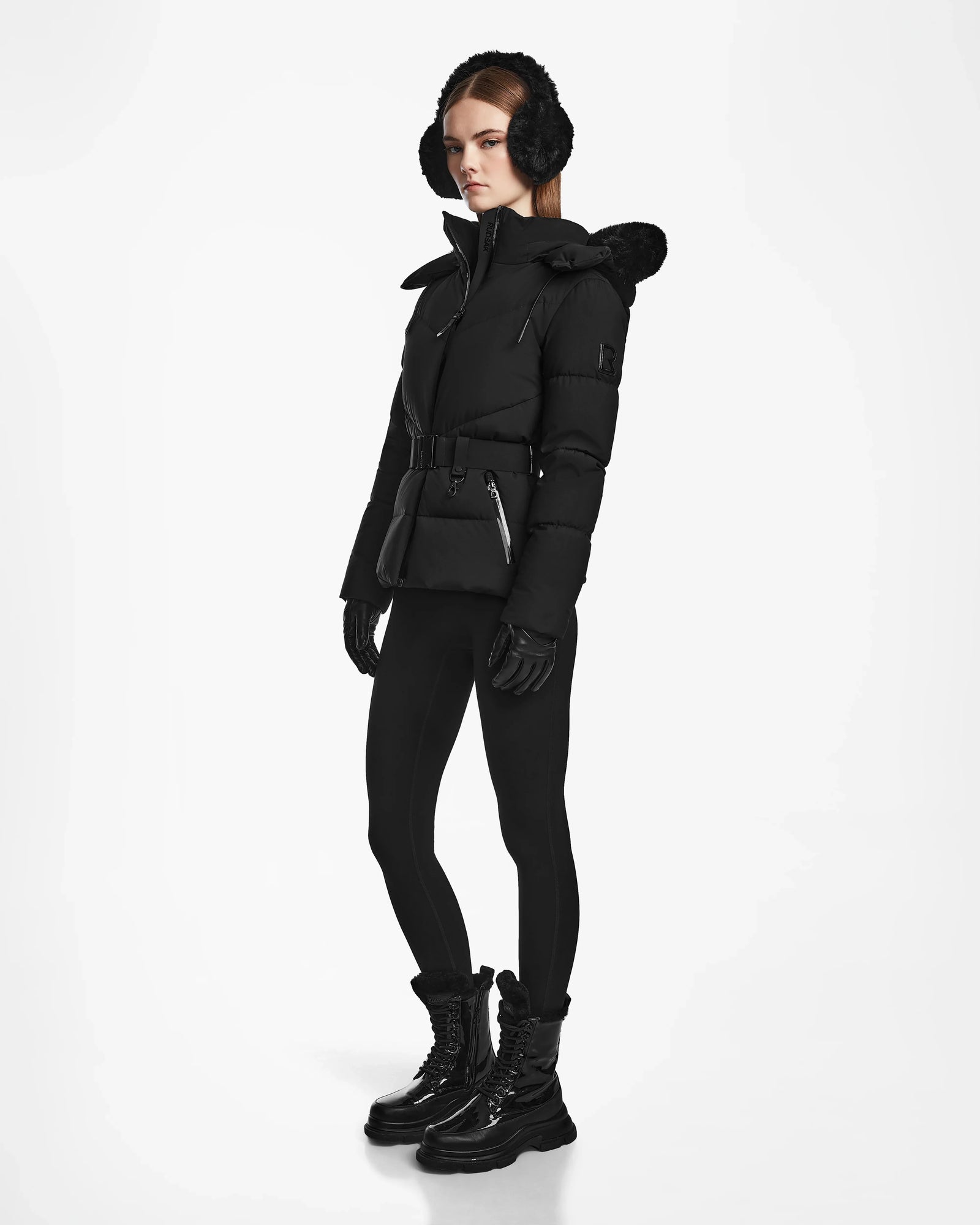 A woman with straight brown hair and a neutral expression models the Rudsak CHRISTELLE M - L123525 Black quilted down puffer, styled with earmuffs, leggings, gloves, and lace-up boots against a white background.