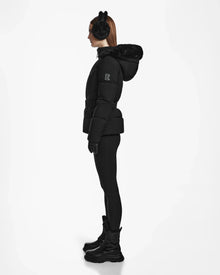 A woman in profile stands against a white background, wearing the Rudsak CHRISTELLE M - L123525 Black sustainable winter coat with a belted, fitted silhouette and fur-trimmed hood, paired with black earmuffs, leggings, gloves, and lace-up boots.