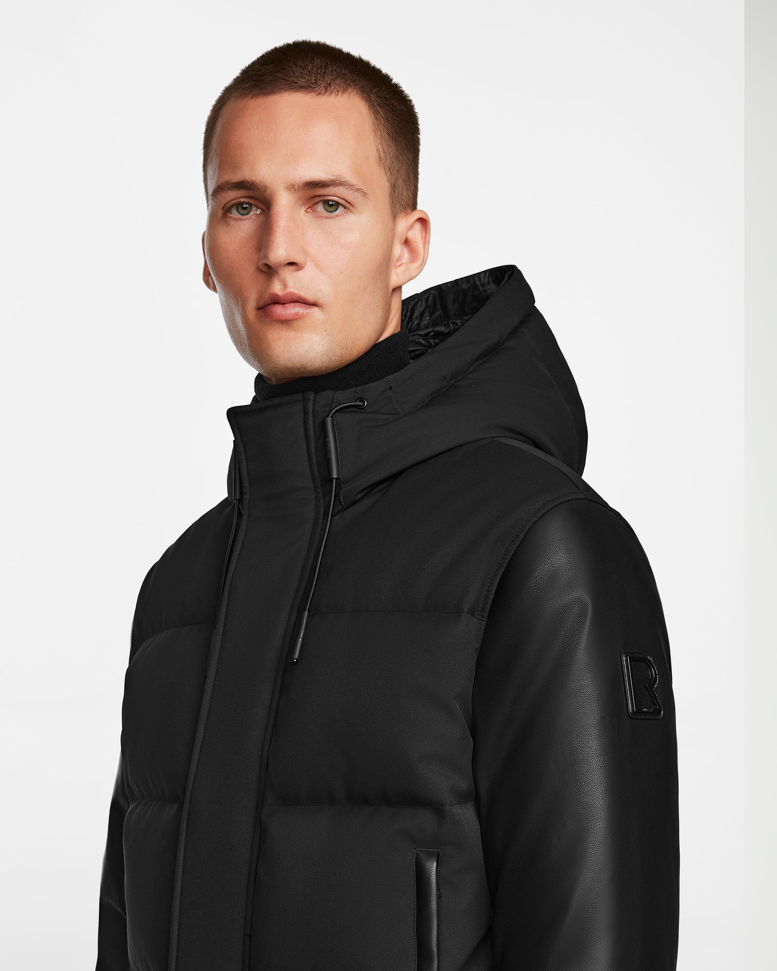 A man with short hair models the Rudsak DALLAS R - M123501 BLACK quilted down bomber with recycled leather sleeves, looking at the camera against a plain white background.