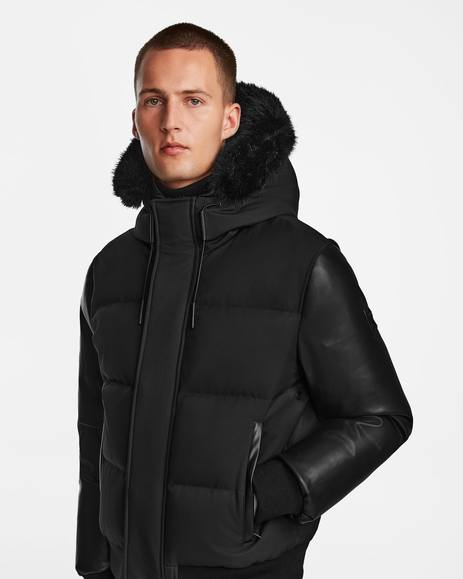 A person models the Rudsak DALLAS R - M123501 BLACK: a men's black quilted down bomber with recycled leather sleeves and a fur-lined hood, standing against a white background and looking neutrally at the camera.