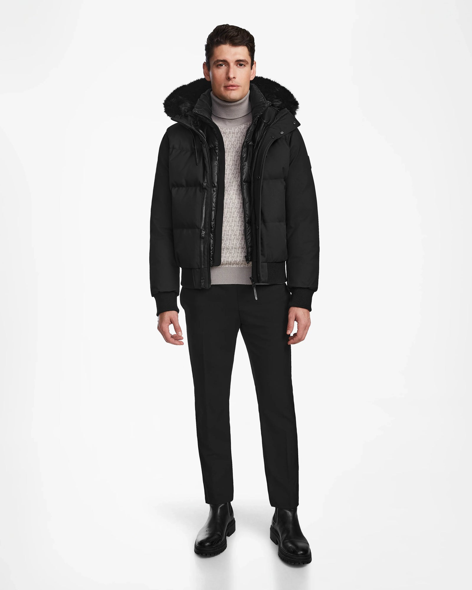 A man stands against a plain white background wearing the Rudsak DALLAS - M122501 Black weather-resistant puffer coat with a fur-lined hood, paired with a light gray turtleneck, black pants, and black dress shoes.